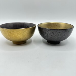 May include: Two ceramic bowls with a metallic finish. One bowl is gold on the inside and dark gray on the outside. The other bowl is dark gray on the inside and gold on the outside. Both bowls have a textured surface.