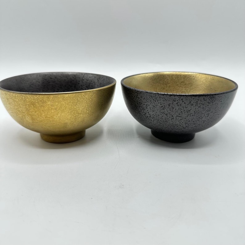 May include: Two ceramic bowls with a metallic finish. One bowl is gold on the inside and dark gray on the outside. The other bowl is dark gray on the inside and gold on the outside. Both bowls have a textured surface.