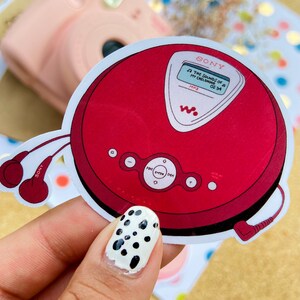 May include: Red and silver Sony CD player with a white screen displaying "THE SOUNDS OF MY CHILDHOOD 02:34 MP3". The CD player has a black headphone jack and a black cord.