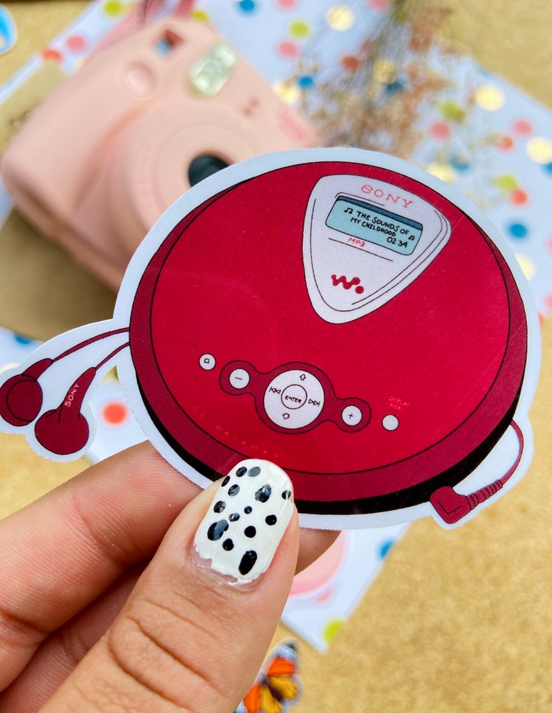 May include: Red and silver Sony CD player with a white screen displaying "THE SOUNDS OF MY CHILDHOOD 02:34 MP3". The CD player has a black headphone jack and a black cord.