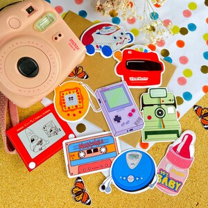 May include: A collection of vintage toy and game themed stickers featuring an instant camera, a View-Master, a Game Boy, a cassette tape, a walkman, a camera, and a toy truck. The stickers are on a brown surface with colorful polka dots.