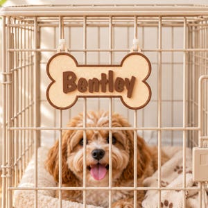 May include: A beige metal dog crate containing a small, fluffy, light brown dog resting on a cream-coloured blanket with brown paw prints. A bone-shaped sign with a brown border and the name Bentley in brown lettering hangs from the crate door.