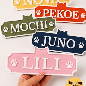 May include: Five rectangular wall signs with cat ear silhouettes, each featuring a white border, white lettering, and white paw prints. The signs are colored yellow, red, olive green, dark blue, and pink, displaying the names NOVA, PEKOE, MOCHI, JUNO, and LILI.