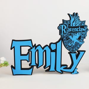Könnte beinhalten: Blau-schwarzes Namensschild mit dem Namen "Emily". Das Schild hat ein Ravenclaw-Wappen. Ein dekorativer Artikel für das Kinderzimmer.