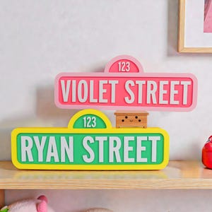 May include: Two vibrant street signs, one pink and one green, with white lettering. The pink sign reads "VIOLET STREET" and the green sign reads "RYAN STREET". Both signs display the number "123".