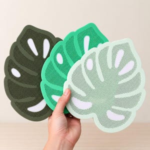 May include: Three leaf-shaped coasters in varying shades of green, including dark olive, grass green, and light mint. Each coaster features a perforated texture and white cut-out details mimicking the natural fenestrations of a monstera leaf.