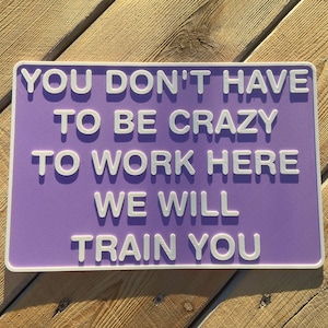 May include: A rectangular purple sign with a white border and raised white lettering that reads: YOU DON'T HAVE TO BE CRAZY TO WORK HERE WE WILL TRAIN YOU. The sign features a smooth, matte finish and is presented in a clear, bold, sans-serif font.