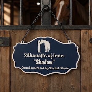 May include: A navy blue, scalloped-edge sign with a white silhouette of a person and a horse. The text reads: Silhouette of love. "Shadow" Owned and Loved by Rachel Moore. The sign hangs from a black metal chain against a wooden stall door.