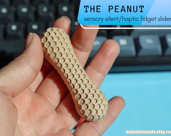May include: A beige, peanut-shaped fidget slider with a textured surface. The slider has a honeycomb pattern and is held in a hand. Text at the top reads "THE PEANUT sensory silent/haptic fidget slider". The background includes a keyboard and a blue surface.