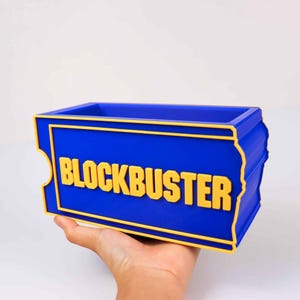 Op de afbeelding: Een rechthoekige blauwe opbergbak met een gele rand en het woord BLOCKBUSTER in verhoogde gele letters aan de voorzijde. De bak heeft een ticketvormig silhouet met ingekeepte zijkanten en een open bovenkant voor het opbergen van items.