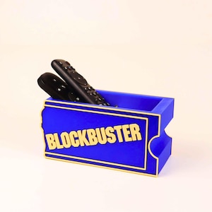 Op de afbeelding: Een rechthoekige blauwe opbergbak met gele rand en het woord BLOCKBUSTER in gele letters op de voorzijde. De bak heeft de vorm van een ticket en bevat twee zwarte afstandsbedieningen in het open compartiment aan de bovenzijde.