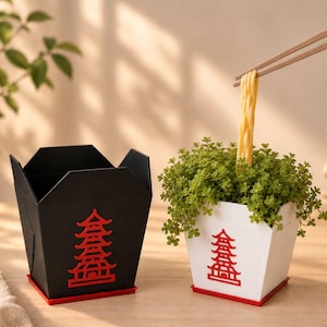 Puede incluir: Dos recipientes cuadrados con forma de caja de comida para llevar, uno negro y otro blanco, cada uno con un gráfico de pagoda roja en el frente y una base roja. El recipiente blanco contiene una planta verde con palillos marrones levantando un fideo.