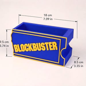 Op de afbeelding: Een blauwe rechthoekige bureau-organizer met het gele Blockbuster-logo op de voorzijde. De afmetingen zijn 18 cm lang, 9,5 cm hoog en 8,5 cm diep. Het ontwerp bootst een klassiek filmverhuurticket na met opstaande gele randen en letters.