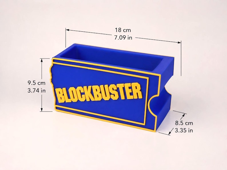 Op de afbeelding: Een blauwe rechthoekige bureau-organizer met het gele Blockbuster-logo op de voorzijde. De afmetingen zijn 18 cm lang, 9,5 cm hoog en 8,5 cm diep. Het ontwerp bootst een klassiek filmverhuurticket na met opstaande gele randen en letters.