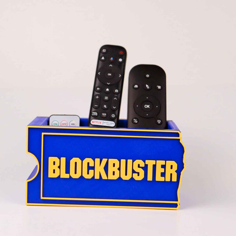 Op de afbeelding: Een blauwe rechthoekige afstandsbedieningshouder met het gele Blockbuster-logo op de voorzijde. De houder bevat twee zwarte televisie-afstandsbedieningen en &eacute;&eacute;n kleine witte afstandsbediening met diverse knoppen, die er rechtop in staan.