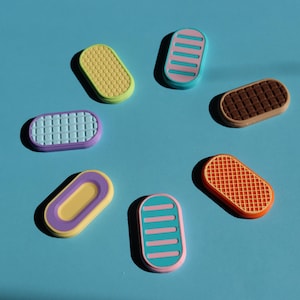May include: A selection of colourful, oval-shaped decorative magnets. The magnets display various designs, including textured patterns, stripes, and grid-like arrangements. The colour palette includes yellow, pink, purple, brown, and orange, set against a blue background.