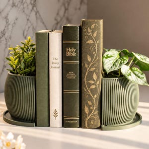 Peut inclure: Deux jardinières vert olive, semi-circulaires et nervurées avec plateaux assortis, placées de chaque côté de quatre livres. Les livres comprennent un journal vert, un journal blanc et deux livres vert foncé, dont une Bible et un livre à motifs dorés.