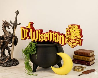 May include: A custom red and yellow sign reading Dr. Wiseman with a Gryffindor crest, a bronze dragon statue with a sword, a black cauldron with green foliage, a yellow crescent moon light, three stacked books, a compass, and a quill pen.