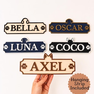 May include: Five rectangular wall signs with decorative edges, each featuring a pet name: BELLA, OSCAR, LUNA, COCO, and AXEL. Each sign includes a silhouette of dog ears or a head at the top center and two paw prints on the sides. Text reads Hanging Strip Included.