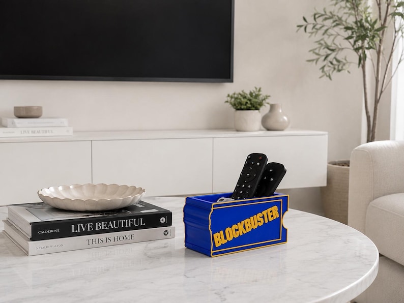 Op de afbeelding: Een rechthoekige blauwe opbergbak met gele rand en het woord BLOCKBUSTER in het geel op de voorzijde gedrukt. De bak bevat twee zwarte afstandsbedieningen en staat op een wit marmeren oppervlak naast een stapel boeken.