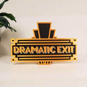 May include: A rectangular sign with a black background and gold-colored raised lettering that reads DRAMATIC EXIT. The sign features a geometric border and a central top crest, all rendered in a bold, layered, and three-dimensional design.