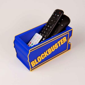 Op de afbeelding: Een blauwe rechthoekige opbergbak met gele rand en het woord BLOCKBUSTER in gele letters in reli&euml;f op de voorzijde. De bak bevat twee zwarte afstandsbedieningen en &eacute;&eacute;n kleine witte afstandsbediening met gekleurde knoppen erin.