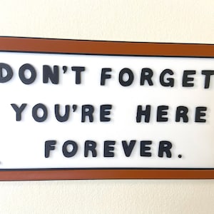 May include: A rectangular wall sign with a brown frame and a white background. The sign features raised black lettering that reads: DON'T FORGET: YOU'RE HERE FOREVER. The text is arranged in three lines, secured by small black corner fasteners.