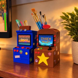 May include: Two arcade-style desk organizers for pens and brushes. The blue unit displays Junimo Kart on its screen, while the brown unit features a large yellow star and the text Stardew Valley on its screen, both decorated with game stickers.