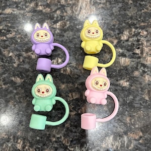 May include: Four bottle stoppers in a cartoon bunny design. Each stopper is a different colour: purple, yellow, green, and pink. They have a loop and a cylindrical base.