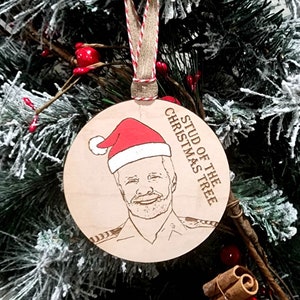May include: A round wooden Christmas ornament with a laser-engraved image of a man wearing a Santa hat. The text "Stud of the Christmas Tree" is engraved on the ornament.