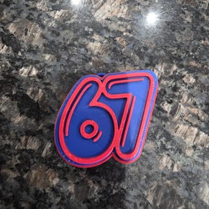 May include: A blue and red number 67, possibly a cookie cutter or decorative piece. The number has a bold, rounded design with a red outline and a blue interior. It rests on a dark, speckled surface.