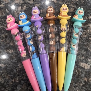 May include: Six colourful pens with animal-shaped toppers. Each pen has a different barrel colour: pink, blue, purple, brown, yellow, and teal. The clear barrels contain small animal figures.