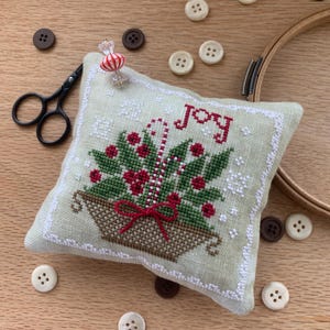 May include: A light green, square pincushion with a cross-stitched design of a basket filled with greenery, red berries, and candy canes. The word "JOY" is stitched in red. A red and white striped pin is inserted. Small buttons and scissors are scattered around.