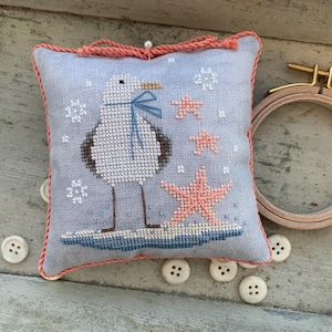 May include: A square pincushion featuring a cross-stitch design of a seagull wearing a blue scarf, standing on a blue patch with two pink starfish and white snowflake-like accents. The cushion is edged with coral-colored twisted cord trim.