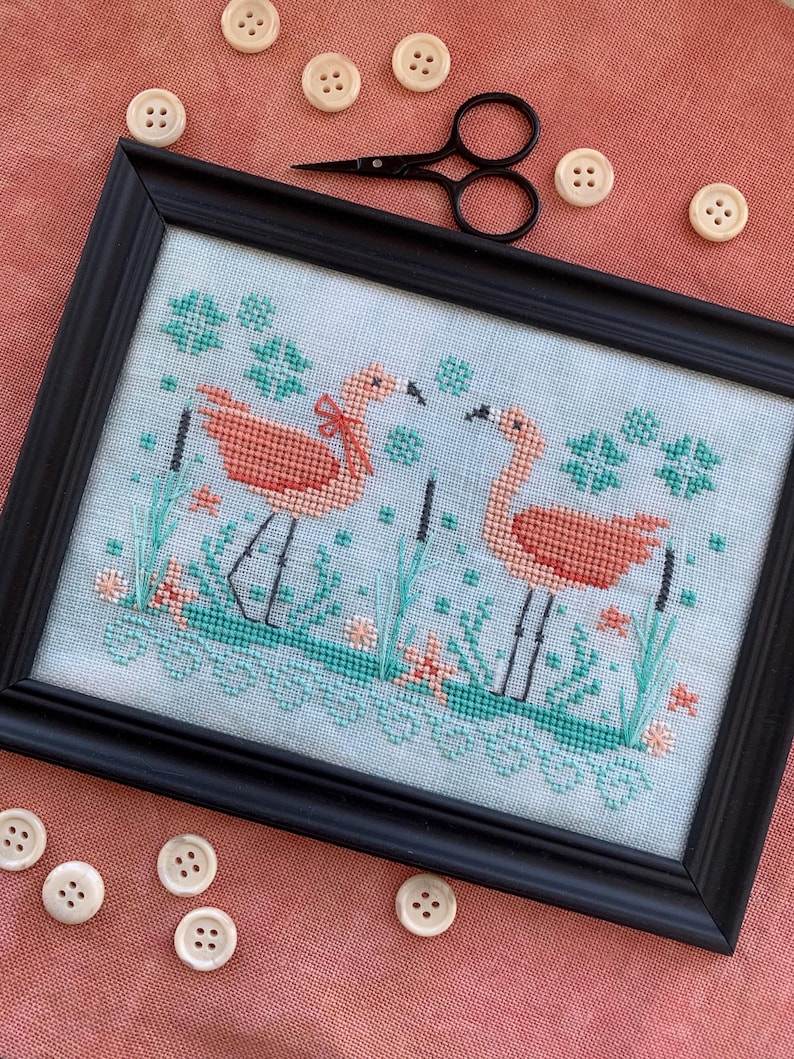 Puede incluir: Arte de punto de cruz enmarcado con dos flamencos rosas con detalles turquesa y coral. La obra est&aacute; en un marco negro, rodeada de botones color crema y unas peque&ntilde;as tijeras negras. El fondo es de tela color coral.