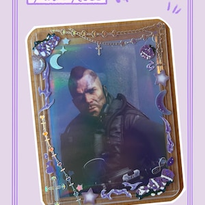 Peut inclure: Un porte-carte rectangulaire en plastique transparent contenant le portrait d'une personne avec une crête. Le support est décoré d'autocollants violets et argentés, notamment des étoiles, des lunes, des croix et des chaînes. Le texte Dark Rose est visible.