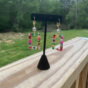 May include: A pair of gold-toned rectangular earrings with colorful sequins and a clear gem at the top. The earrings are displayed on a black stand against a blurred green background.