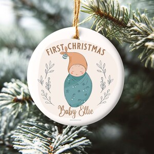 May include: A white ceramic Christmas ornament with a brown and blue illustration of a baby swaddled in a blanket. The ornament reads "First Christmas Baby Ellie".