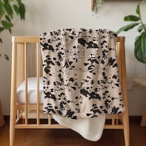 May include: A beige and black cow print blanket with the name "ELLIE MAE" in a rope-style border. The blanket is draped over a wooden crib, with a white fluffy underside visible.