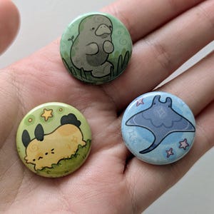 May include: Three round, colorful pinback buttons featuring cute animal illustrations. One button shows a gray manatee, another a yellow and black creature, and the last a blue manta ray with stars. The buttons are held in a hand.