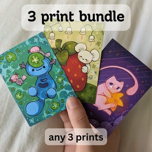 Puede incluir: Un conjunto de tres impresiones artísticas con ilustraciones coloridas. Una impresión muestra un personaje de dibujos animados azul, otra un ratón con una fresa y la última un gato rosa con una estrella. El texto dice "3 print bundle" y "any 3 prints".