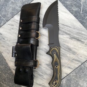 Könnte beinhalten: Ein schwarzes Überlebensmesser mit gezahnter Klinge und Holzgriff mit Messingnieten. Das Messer liegt neben einer schwarzen Lederscheide mit Messingringen und Riemen.