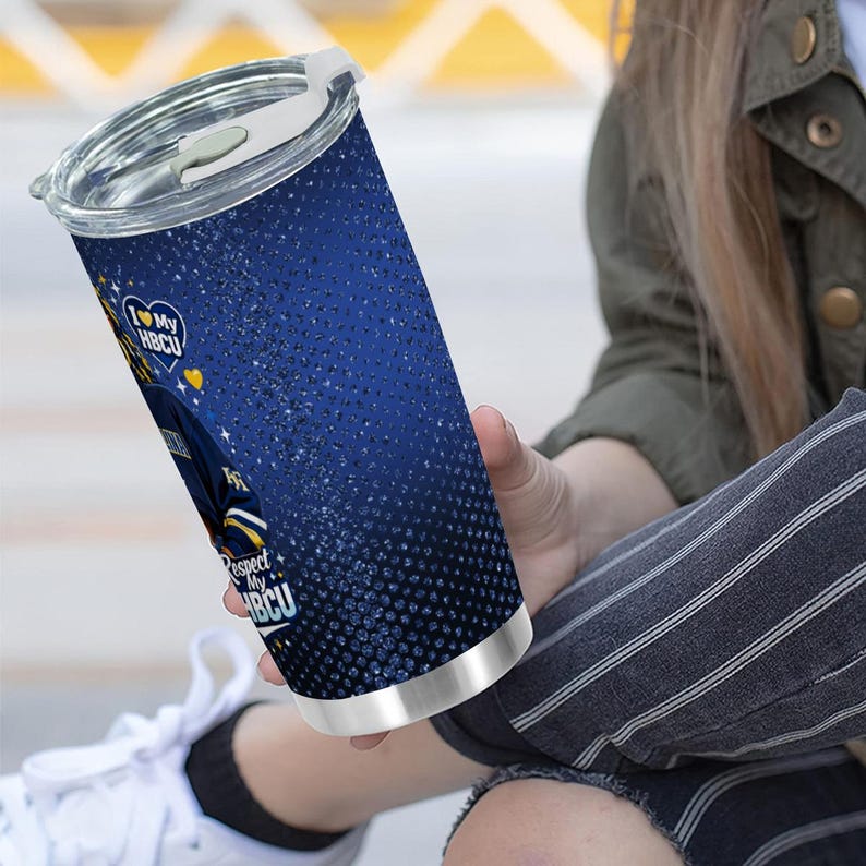 May include: A blue and silver insulated tumbler with a lid. The tumbler features the text "I love my HBCU" and "Respect my HBCU" with a design of a person in a jacket. The tumbler is held by a person.