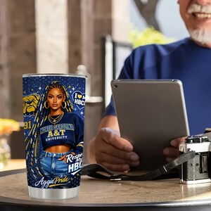 May include: A stainless steel tumbler featuring a graphic of a woman with long braids, wearing a North Carolina A&T University sweatshirt. The tumbler has the text "Aggie Pride" and "Respect My HBCU". The design includes a bulldog logo and the number 91.