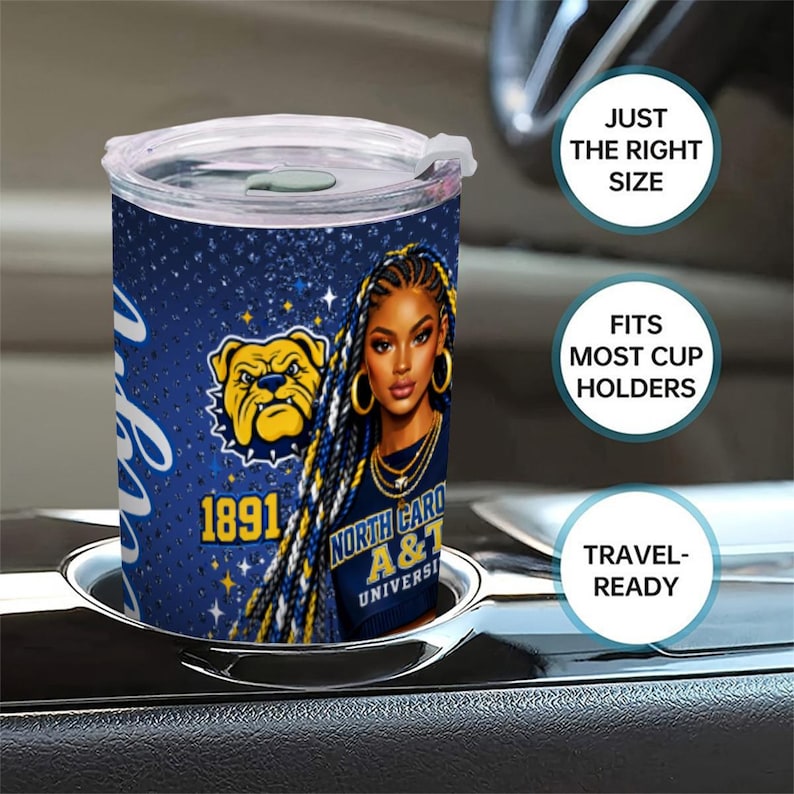 May include: A blue and white travel mug featuring a portrait of a woman with long braids and gold jewelry. The mug also displays the text "North Carolina A&T University" and a bulldog logo. The mug is in a car cup holder.