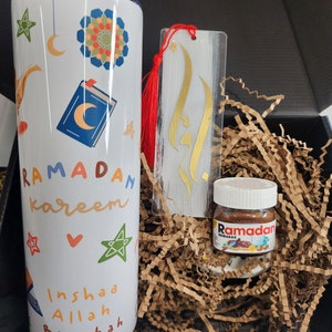 May include: A gift box with a white tumbler, a bookmark, and a small jar of Nutella. The tumbler is decorated with colorful Ramadan-themed illustrations and text. The bookmark has gold calligraphy and a red tassel. The jar of Nutella has a Ramadan label.