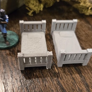 May include: Two miniature gray beds with headboards and footboards. Each bed has a pillow and a blanket. A miniature figurine of a person in blue armor is on the left. The beds are on a wooden surface.
