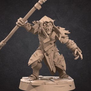 May include: A miniature goblin figurine holding a spear, wearing armour and a hooded cloak. The figurine is painted in a light brown colour and is standing on a circular base.