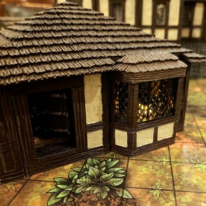 May include: A miniature, detailed model of a rustic building with a brown shingled roof and dark wooden trim. The building has a window with a lattice design and a small open doorway. Green foliage is at the base.