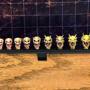 May include: A set of twelve miniature sculpted heads arranged in a row on a black display stand. The heads transition from a pale, flesh-toned human-like appearance on the left to increasingly demonic, yellow-coloured, horned forms on the right.